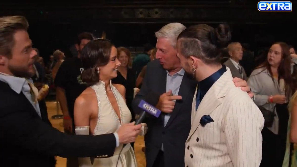 An 'EXTRA' entertainment news interview shows a man with a microphone speaking with a group of people, including a woman in a white dress and a man in a striped suit, at what appears to be a formal event.