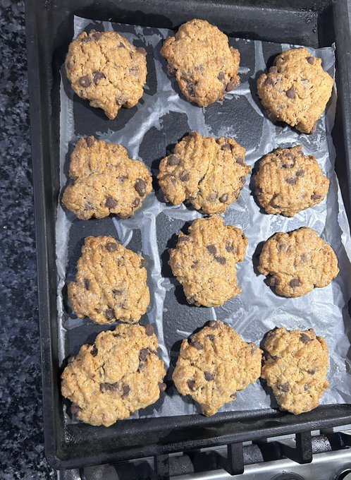 Twelve freshly baked oatmeal chocolate chip cookies are arranged on a parchment-lined baking sheet on a dark countertop.