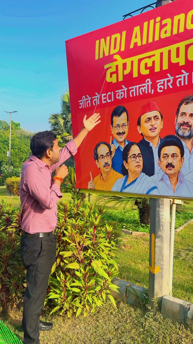 A man in a pink shirt stands with his back to the camera, pointing at a large red billboard for the 'INDIA ALLIANCE' party featuring portraits of several Indian political leaders, including Rahul Gandhi and Akhilesh Yadav.