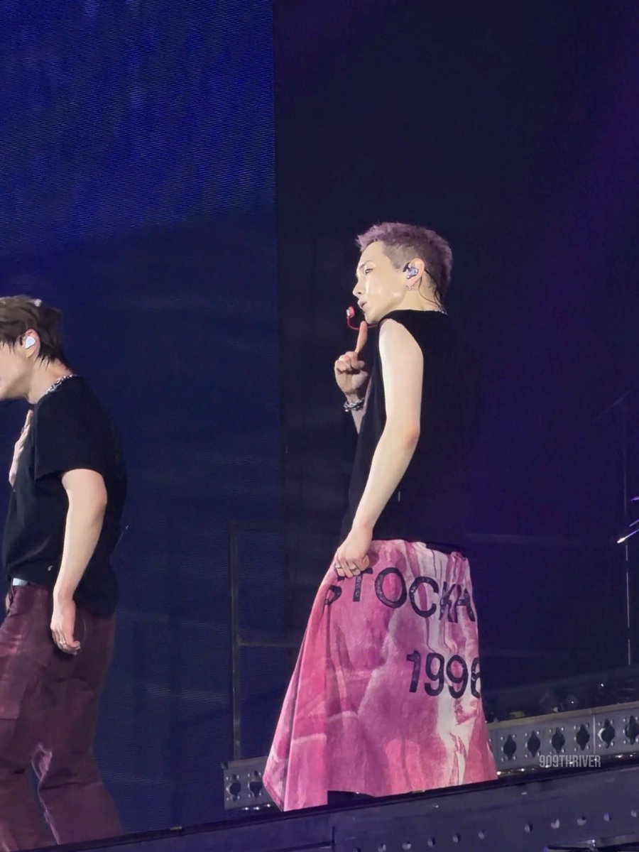 A concert photograph showing a K-pop performer with short purple hair wearing a black sleeveless top and a pink skirt displaying "1998" and "DO99THRIVER" watermark, performing alongside another member in a black shirt.