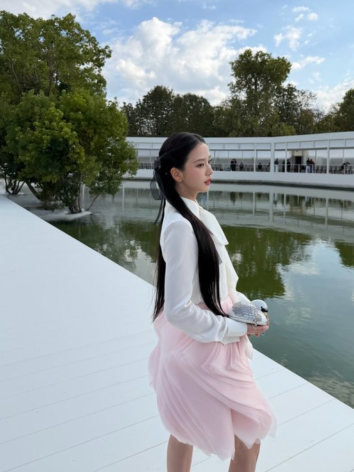 A young woman with long dark hair stands on a white deck beside a pond, wearing a white blouse and pink skirt.