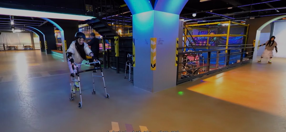 Indoor roller skating rink with a person wearing a helmet and using a walker, alongside another person on roller skates, illuminated by colorful neon and blue lighting.