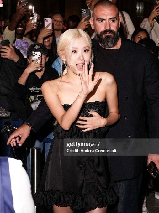 K-pop artist Rosé (Park Chaeyoung) in a black strapless ruffled dress poses with a surprised expression beside a bearded man in black, surrounded by fans taking photos, with watermark 'Credit: Andrea Cremascoli'.
