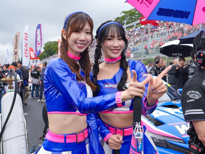Two smiling young women in matching blue and pink WAKO'S racing promotional outfits pose together at what appears to be a motorsport event, with race cars and crowds visible in the background.