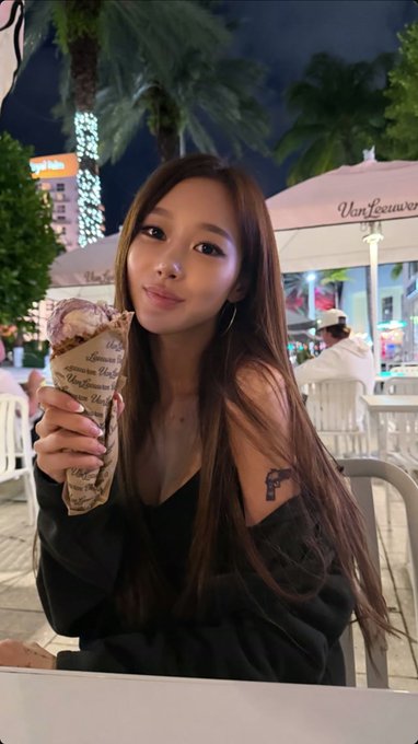 A young woman with long brown hair holds a crepe outdoors at night in front of a 'Van Leeuwen' ice cream shop, wearing a black off-shoulder top.