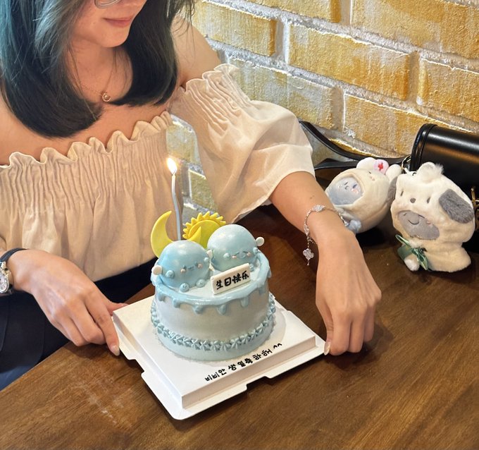 A woman celebrating her birthday with a small light blue cake featuring a lit candle and a moon decoration, with the name '椎名林檎' written on it, sitting at a table next to some plush toys.