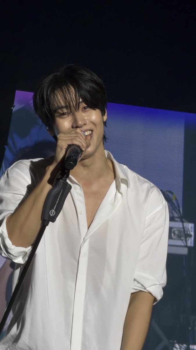 A man wearing an unbuttoned white shirt holds a microphone and smiles while performing on stage.
