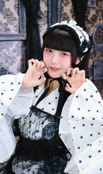 A young woman dressed in a black and white maid-style costume with a polka-dot top and frilly headband poses playfully with her hands near her face.
