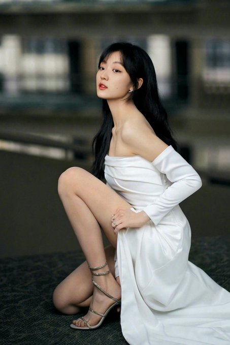A woman with long black hair poses in a white off-the-shoulder dress and strappy heels.