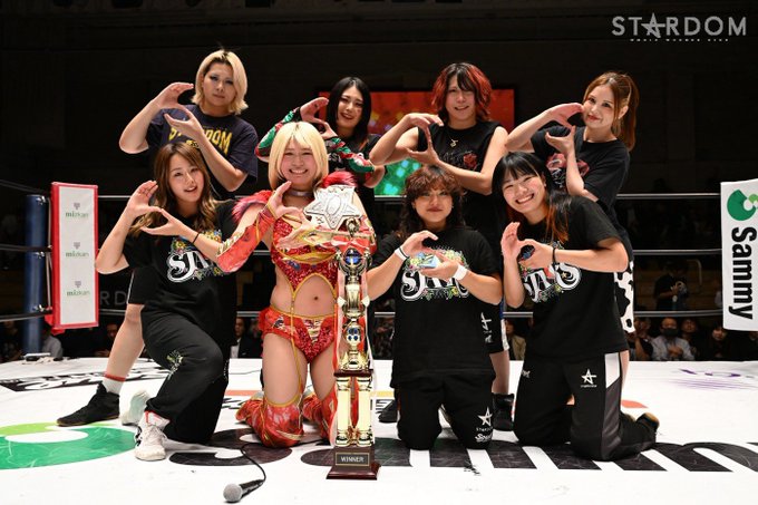 A group of female wrestlers and staff pose for a championship photo with the Stardom (World Wonder Ring Stardom) trophy, making heart shapes with their hands.