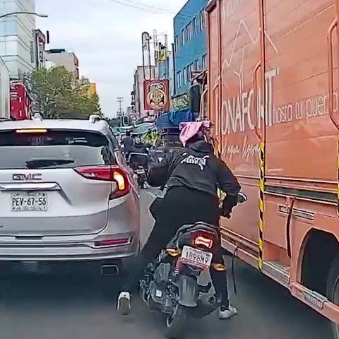 A motorcyclist navigates through congested traffic between a white GMC SUV and an orange bus, with a license plate reading '189GNZ'.