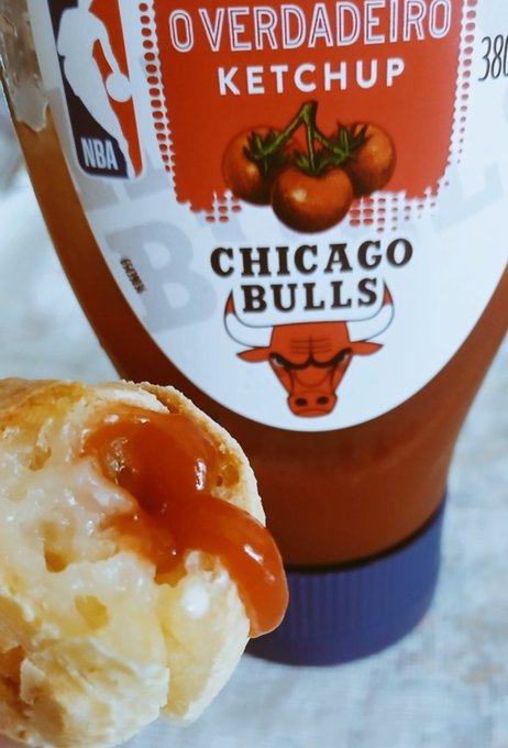 A bottle of 'Overdadeiro Ketchup' featuring Chicago Bulls and NBA branding sits behind a bread roll topped with ketchup and cheese.