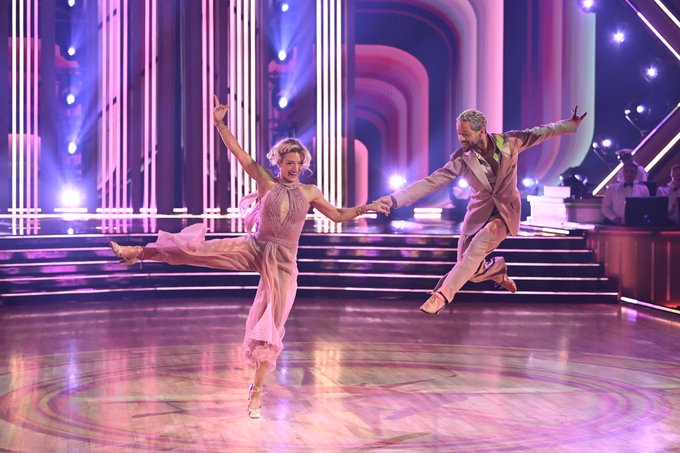 A male and female dancer perform a foxtrot routine mid-leap on a brightly lit stage, holding hands with arms extended gracefully outward. The scene captures a dynamic moment from "Dancing with the Stars" (#DWTS), aligning with the caption about Amber Glenn and Pasha Pashkov's performance during Yacht Rock Night.