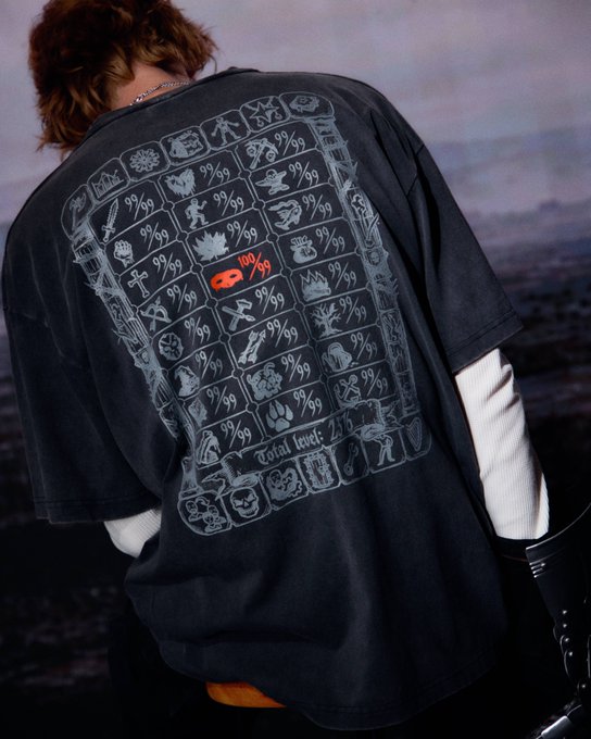 A person models an oversized black t-shirt featuring a gridded back print of gaming and pop-culture icons, each labeled "99/99" in pixel font—with one icon highlighted in red as "100/99" and "total level: 235" printed below, directly echoing the post's "100/99" reference.