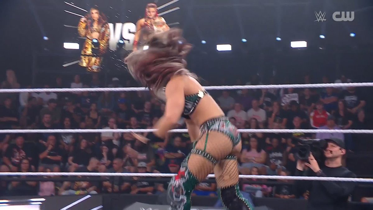 A female WWE wrestler in green gear whips her hair mid-action in a wrestling ring during a televised match, with a "VS" graphic displaying her opponent on the jumbotron above an engaged crowd. The dynamic motion and athletic pose capture the high-energy moment referenced in the post highlighting @ZariaWWE_'s impressive move.