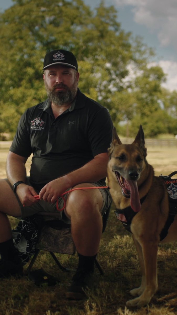 A bearded man wearing a black cap and polo shirt sits outdoors next to a German Shepherd dog wearing a tactical harness.