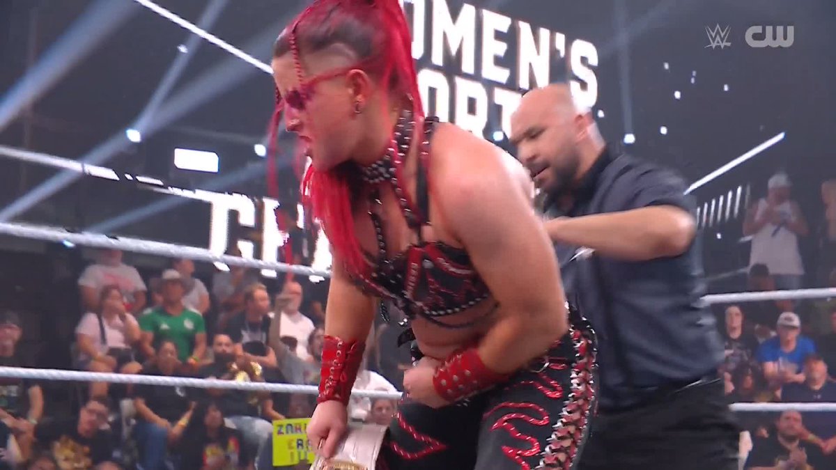 A wrestler with red hair and red-and-black gear stands in the ring holding a championship belt, while a man in a black shirt stands behind her, with "WOMEN'S" visible on the arena signage and the WWE/CW logo in the corner. The image captures the victorious moment referenced in the post, showing Zaria celebrating her title retention in front of an enthusiastic crowd.