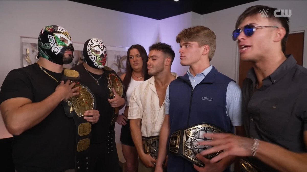 Six individuals stand together backstage, with two masked wrestlers on the left holding championship belts while four unmasked people on the right also display championship titles, exuding the confidence referenced in the caption.