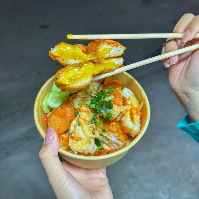 Hands use wooden chopsticks to hold up pieces of fried bread coated in a bright yellow sauce over a bowl containing rice, sliced carrots, fried items, and chopped green onions in an orange sauce.