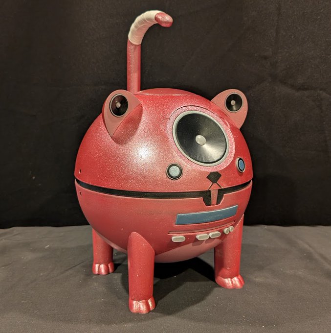A red, spherical robotic cat-like figure with four short legs, frog-like eyes, a long curling tail, and a large central camera lens stands against a black background.