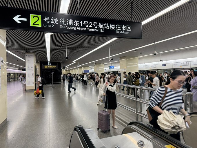 Travelers navigate a busy subway station in China, with a sign reading 'Line 2 To Pudong Airport Terminal 1&2 To Midtown' hanging above the escalators.
