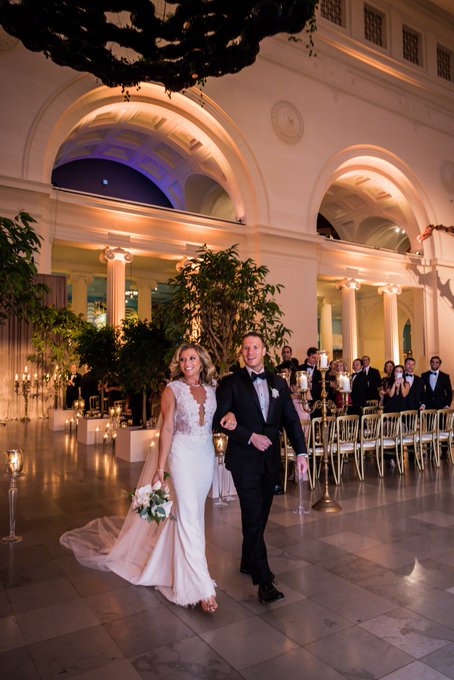 A bride in a white lace gown walks arm-in-arm with a groom in a black tuxedo through an elegant hall with arched columns, while guests seated at candlelit tables applaud.
