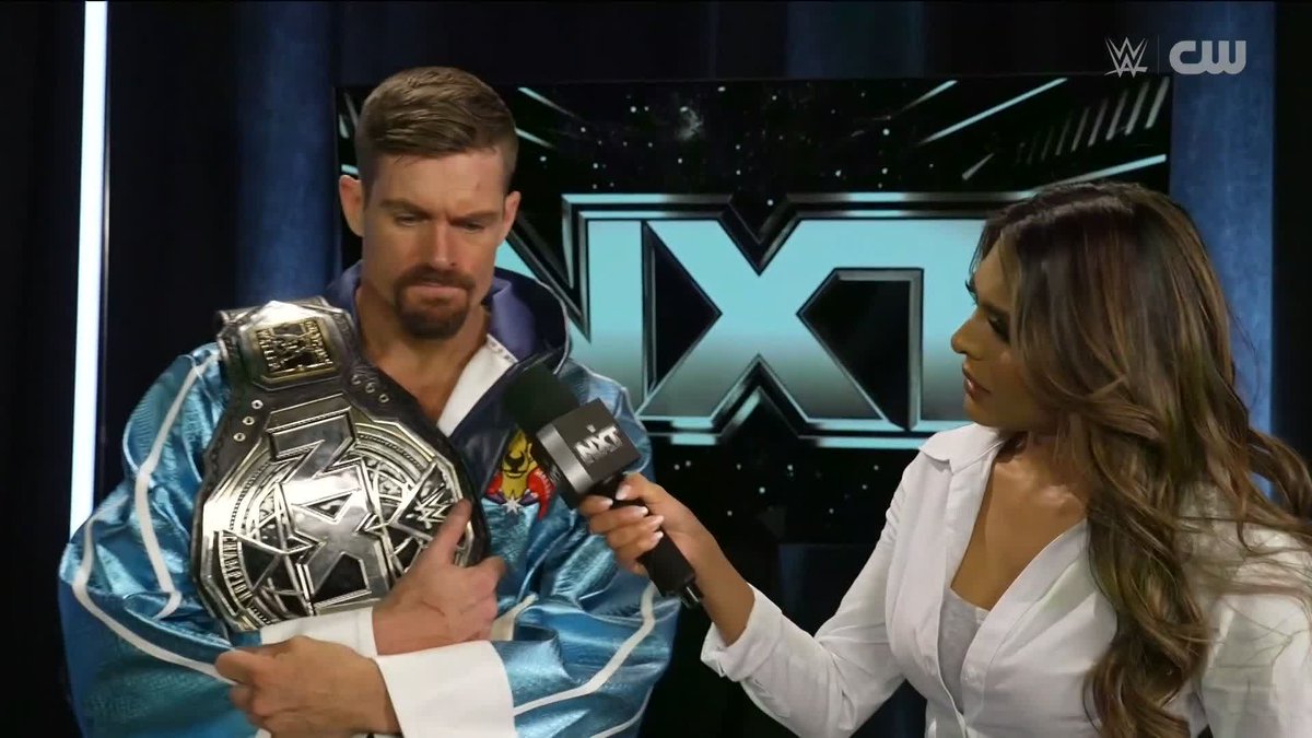 A bearded wrestler in a blue and white jacket holds an NXT championship belt while being interviewed by a woman holding an NXT-branded microphone, in front of a large NXT logo backdrop with WWE and CW network watermarks visible in the corner. The image relates to the post's caption by showing wrestler Grayson Waller delivering a post-match message to the entire NXT Men's locker room while celebrating with his newly won championship title.