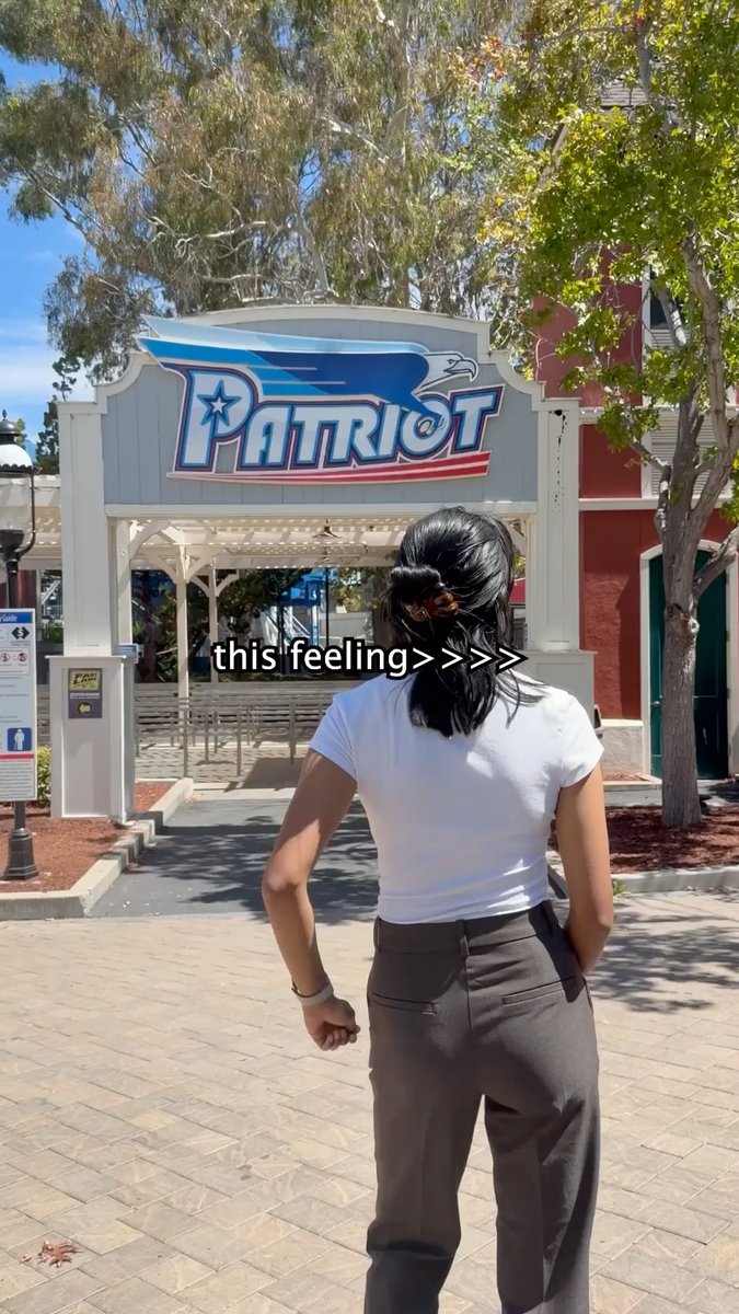 A woman in a white t-shirt and brown pants walks toward the entrance of "Patriot," a wooden roller coaster with red, white, and blue theming featuring an eagle logo. The image captures the anticipation of arriving at a theme park, fitting the caption about enjoying "a few coasters" as a perfect escape.