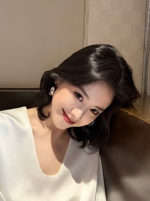 A young woman with shoulder-length dark hair wearing a white top takes a close-up selfie with a soft smile.