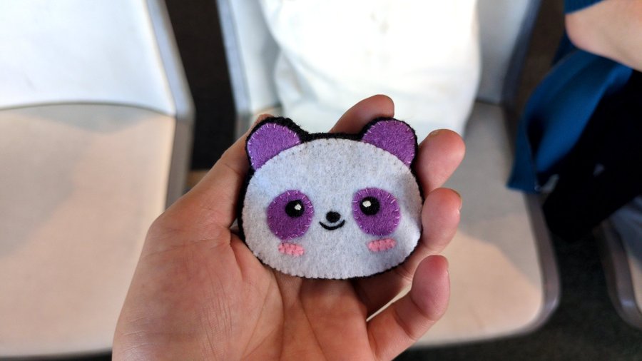 A hand holding a small, round white plush toy with purple embroidered ears, eyes, and cheeks, designed to look like a panda.