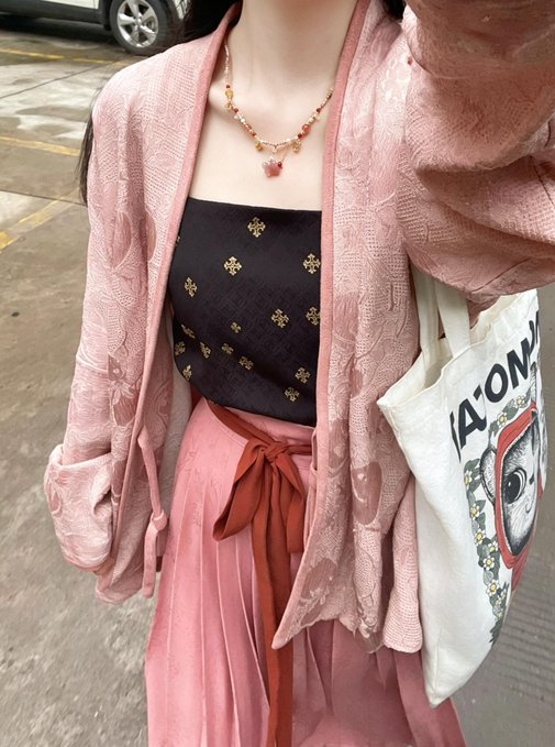 A person wearing a pink knit cardigan over a black tube top with gold patterns and a pink skirt, accessorized with a beaded necklace and carrying a tote bag.