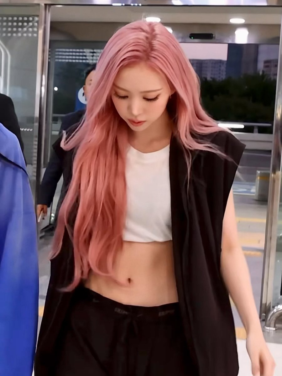 A young woman with long pink hair wearing a white crop top and black sleeveless jacket walks through what appears to be an airport or transit terminal.