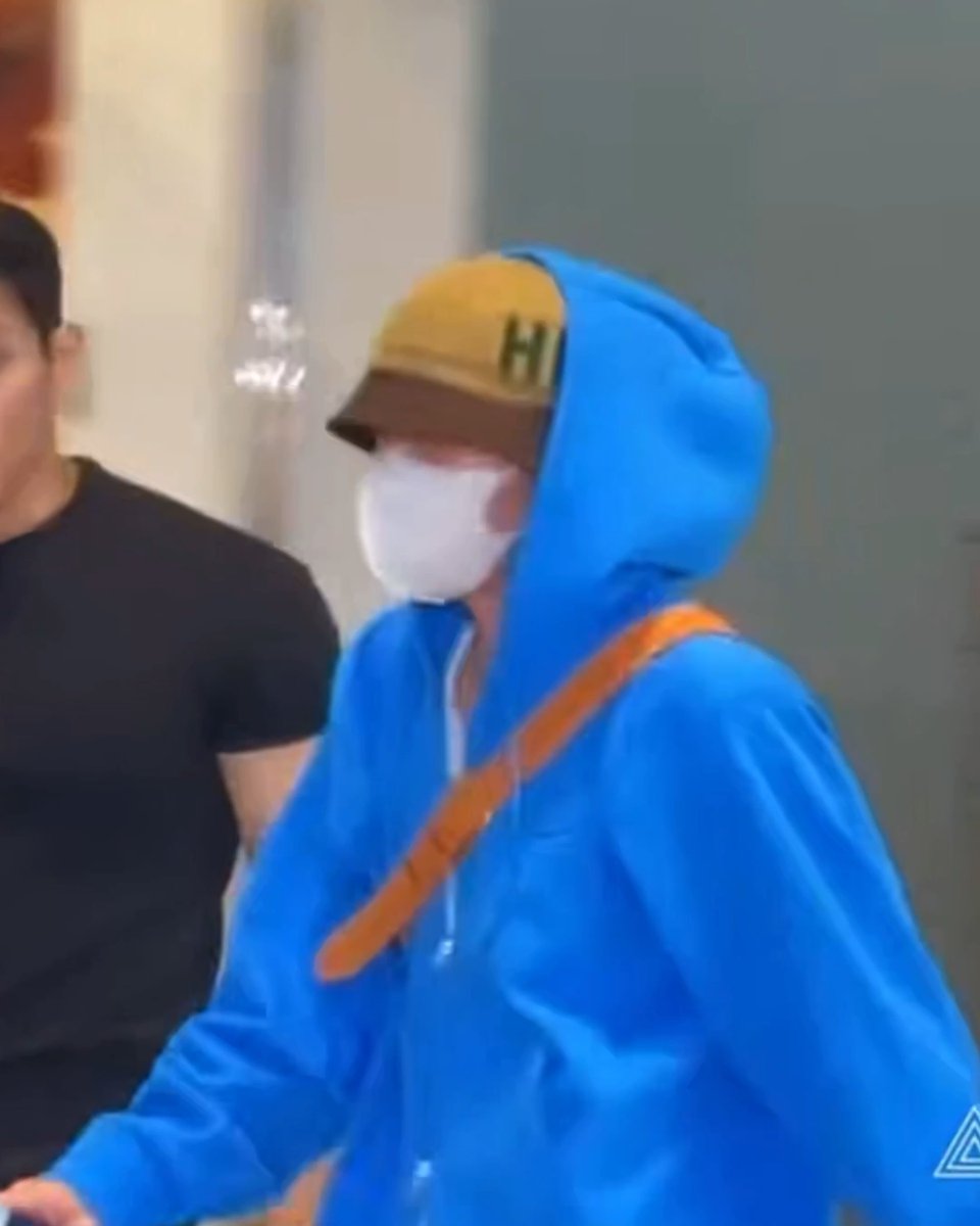 A blurry photo of a person in a blue hoodie, white mask, and yellow cap walking alongside another individual in black.