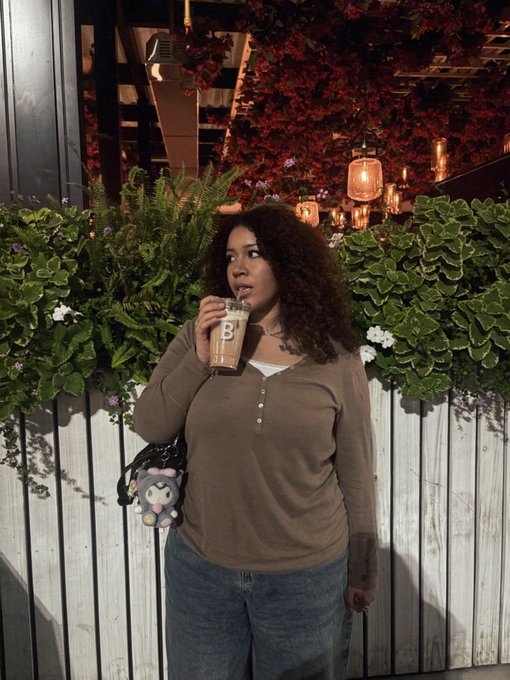 A woman with curly hair drinks from an iced coffee cup with a 'B' logo while standing in front of a white fence decorated with plants and warm pendant lights.