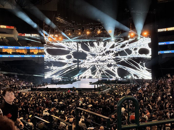 A large indoor arena concert venue with a spectacular stage featuring an X-shaped digital screen display with dramatic white lighting effects, filled with a crowd of spectators.