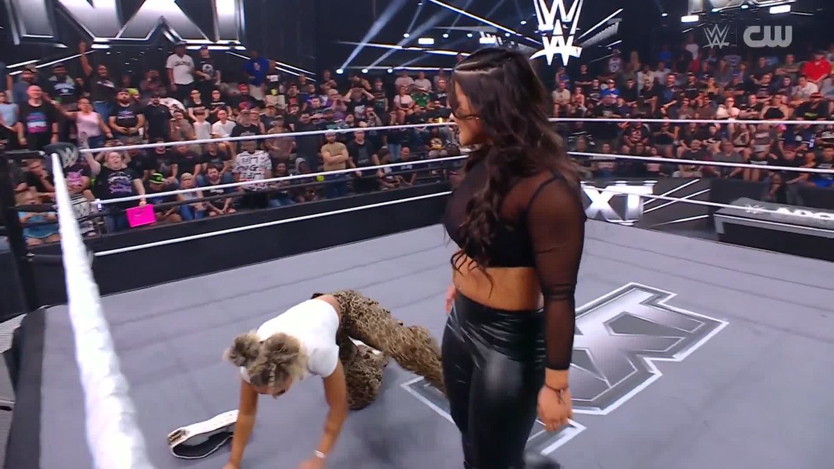 A WWE wrestling match scene on a NXT ring where a female wrestler in black stands over another wrestler who is down on the mat, with a large audience watching in the background.