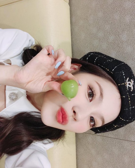 A close-up shows a woman with a black beret, red lipstick, and long blue nails holding a green grape near her face.