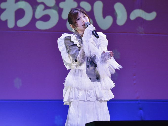 A performer sings into a microphone on stage wearing an elaborate white ruffled dress with lace sleeves, set against a purple backdrop featuring Japanese text.