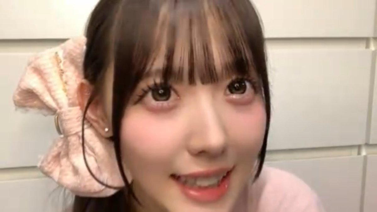 A close-up selfie shows a young woman with dark hair, blunt bangs, and a large pink satin bow smiling softly against a plain tiled wall.