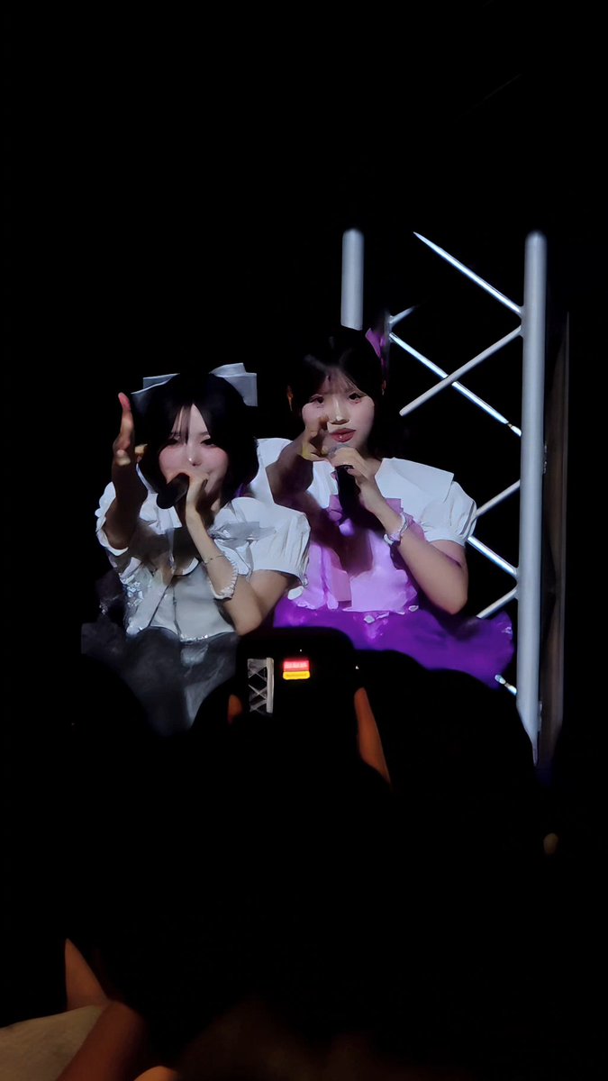 Two young women on a stage with microphones, wearing white and purple outfits with hair bows, appear to be performing live.