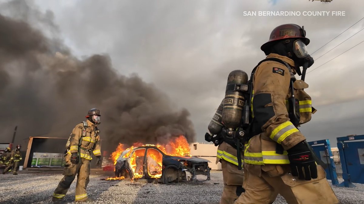 Firefighters in full gear from San Bernardino County Fire responding to a vehicle fully engulfed in flames with thick black smoke billowing into the sky.