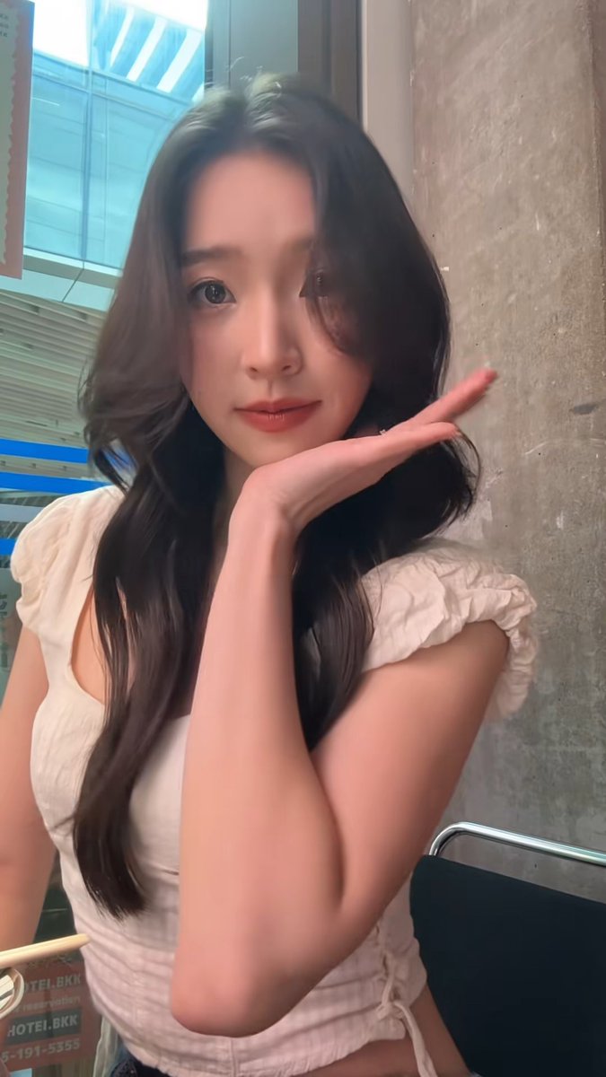A young woman with long dark hair posing with her hand under her chin in front of a concrete wall, with a 'HOTEL BKK' sign partially visible in the background.