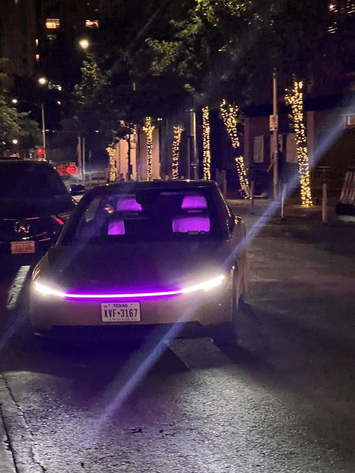 A nighttime photograph of a futuristic Tesla car featuring purple interior lighting and a glowing front light bar, with a Texas license plate reading 'XVF-3167'.