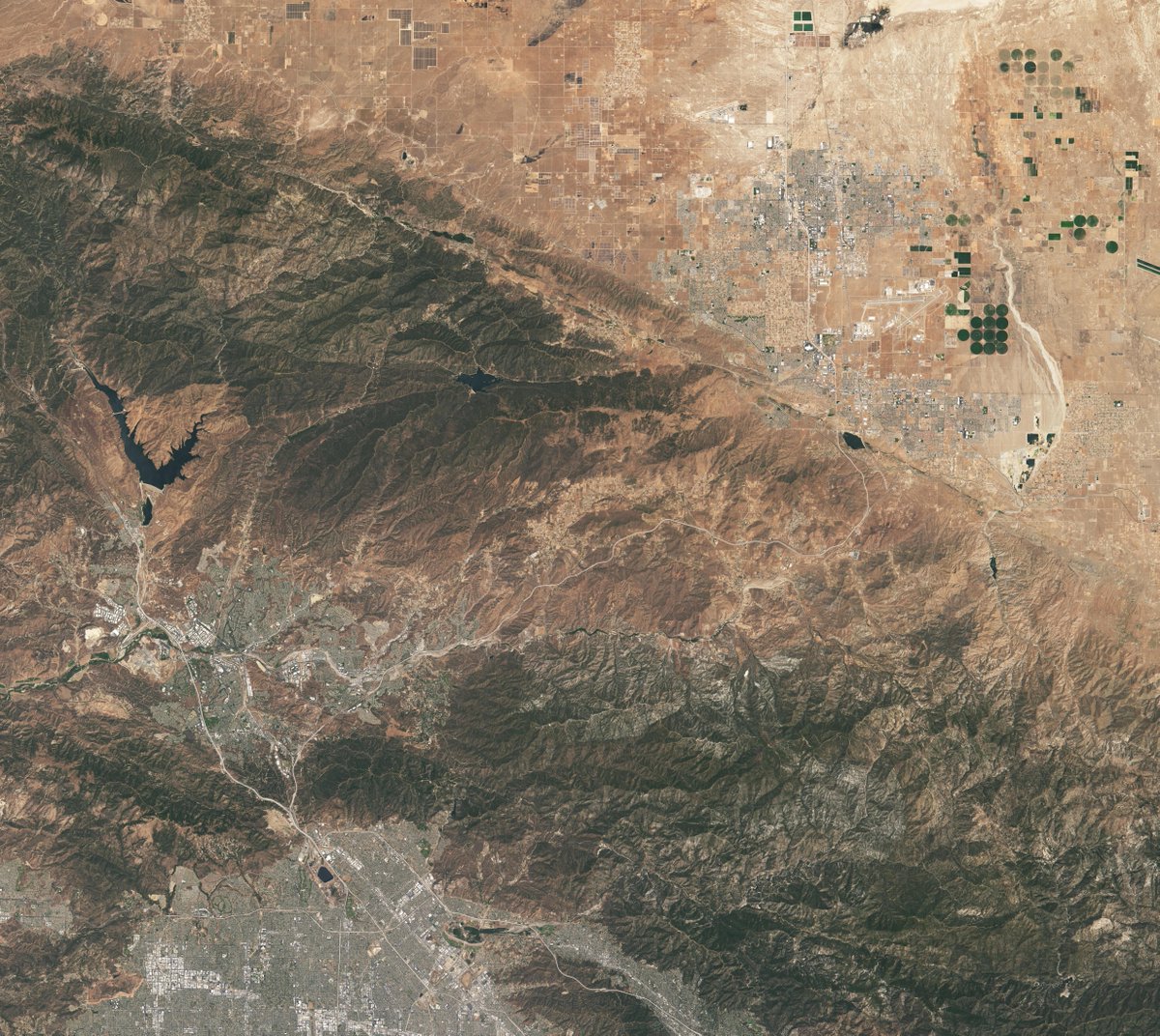 Natural color satellite image showing the Vasquez Rocks Natural Area and surrounding area in Southern California. Most of the landscape is brown and mountainous. The Vasquez Rocks appear as a patch of curved gray stripes close to the bottom left corner near Agua Dulce. Palmdale is visible in the top right corner and the Bouquet Reservoir is near the middle left of the image. 