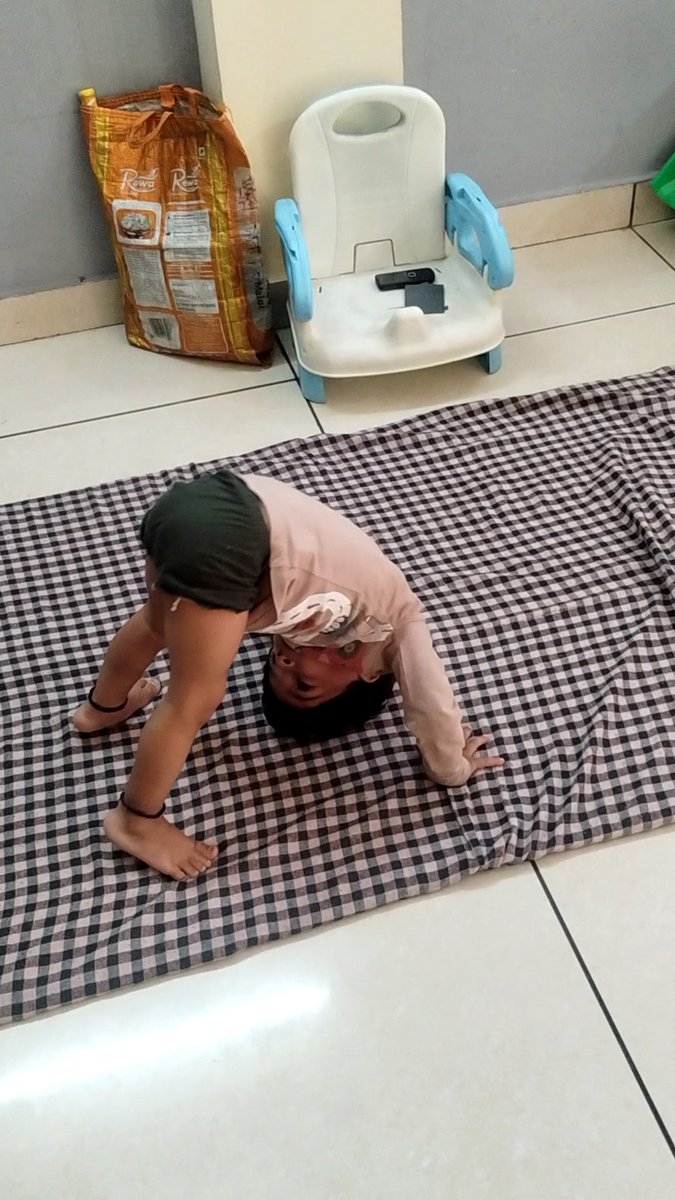 A person performing a backbend or bridge exercise on a black and white checkered mat indoors, with a child's training chair and a large sack visible in the background.