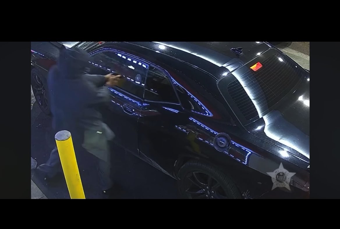 The image shows a blurred surveillance video still of an individual wearing dark clothing and a hood reaching toward a dark-colored vehicle, with a yellow bollard visible in the foreground and a "Sacramento Police" badge watermark in the corner. The footage appears to be from an investigation, though the sensationalized post title does not match what is actually depicted in this crime-related surveillance clip.