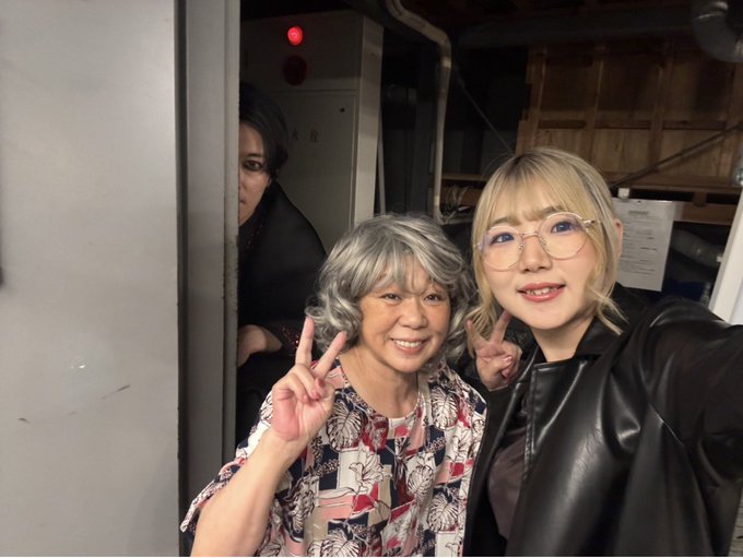 A selfie of two smiling Asian women making peace signs with their fingers—one older with gray curly hair in a floral patterned shirt, and one younger with blonde hair and round glasses wearing a black leather jacket—with another woman partially visible in the background.