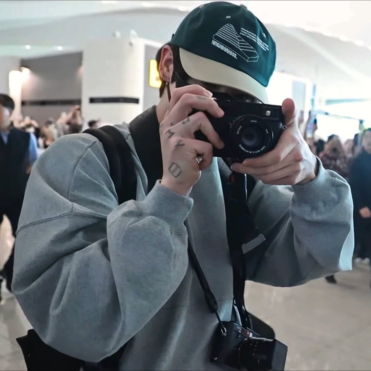 A person wearing a green "NEIGHBORHOOD" cap and gray sweatshirt holds a Leica camera up to their face, with tattooed fingers gripping the device in a crowded indoor setting.