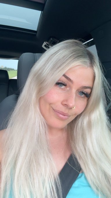 A young woman with long, straight platinum blonde hair and blue eyes smiles at the camera while sitting in the passenger side of a car. She wears a turquoise top, a small nose ring, and a black seatbelt across her chest.