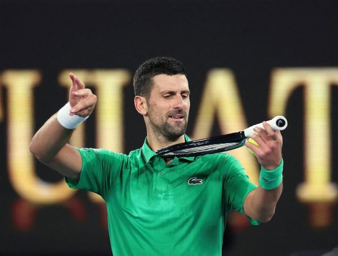 Tennis player Novak Djokovic, wearing a green Lacoste polo, holds a tennis racket and points while holding a tennis ball at what appears to be the Australian Open.